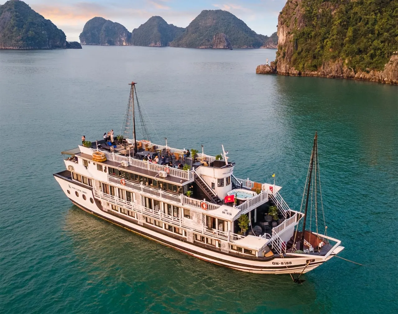 Aerial view of a white multi-deck cruise boat with registration text on the stern and hull, a Vietnamese flag, and people on the top deck, sitting on green water among forested limestone karst islands at sunset.