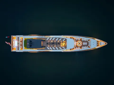 A top-down aerial view at dusk of a large luxury cruise vessel with warm lit decks, lounge chairs and a spiral staircase, floating on dark water with small kayaks or paddleboards near the stern.