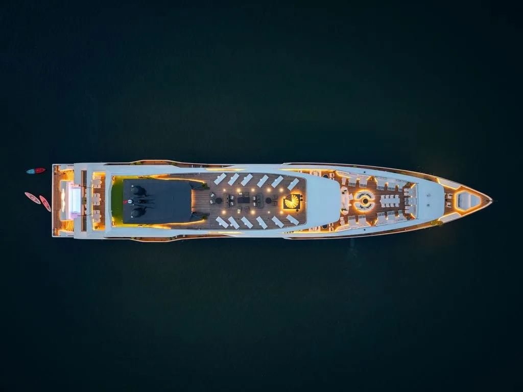A top-down aerial view at dusk of a large luxury cruise vessel with warm lit decks, lounge chairs and a spiral staircase, floating on dark water with small kayaks or paddleboards near the stern.