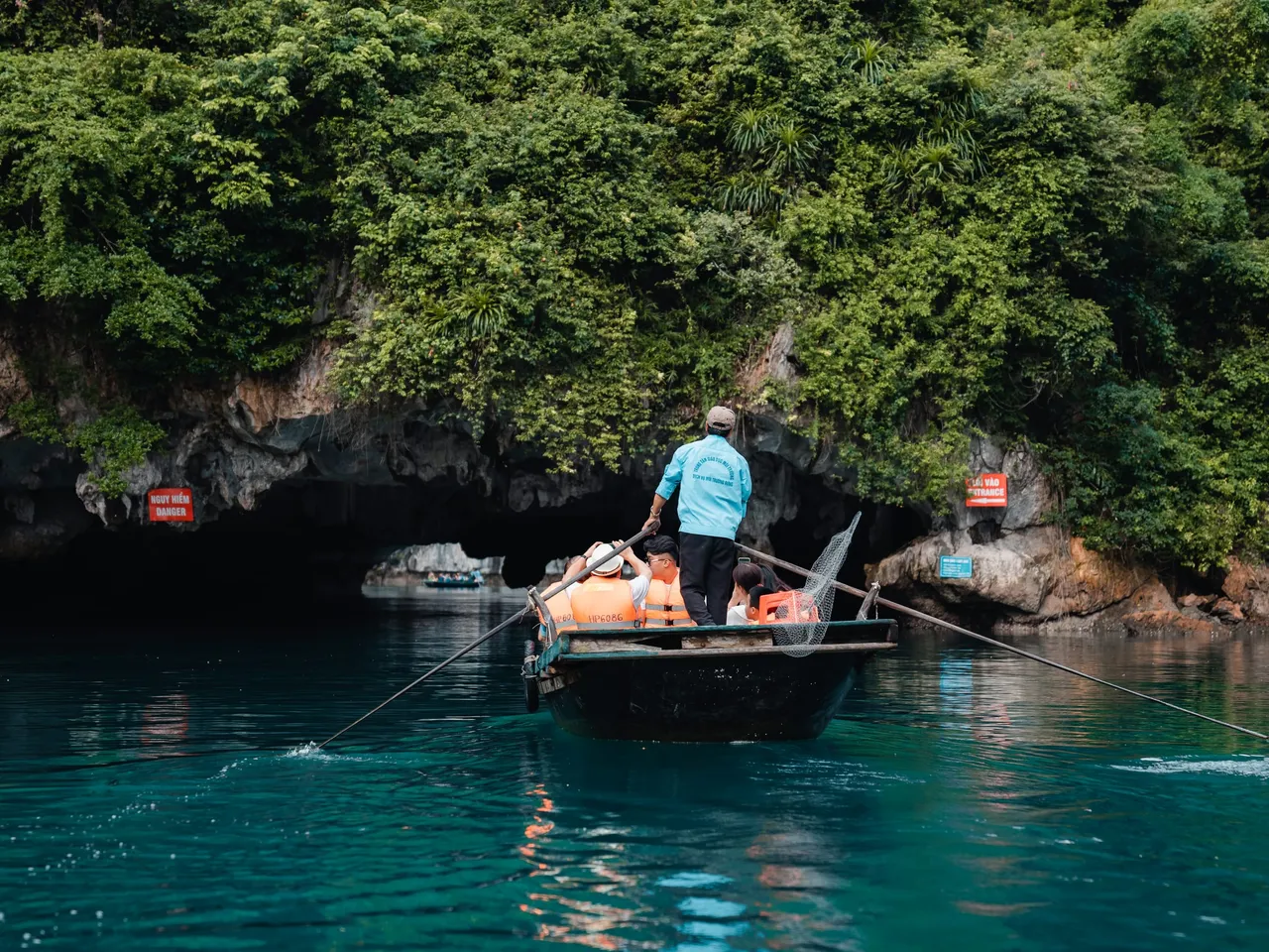 A person in a turquoise shirt rows a small wooden boat with orange life jackets past a cave entrance in green karst cliffs with red signboards mounted on the rock.