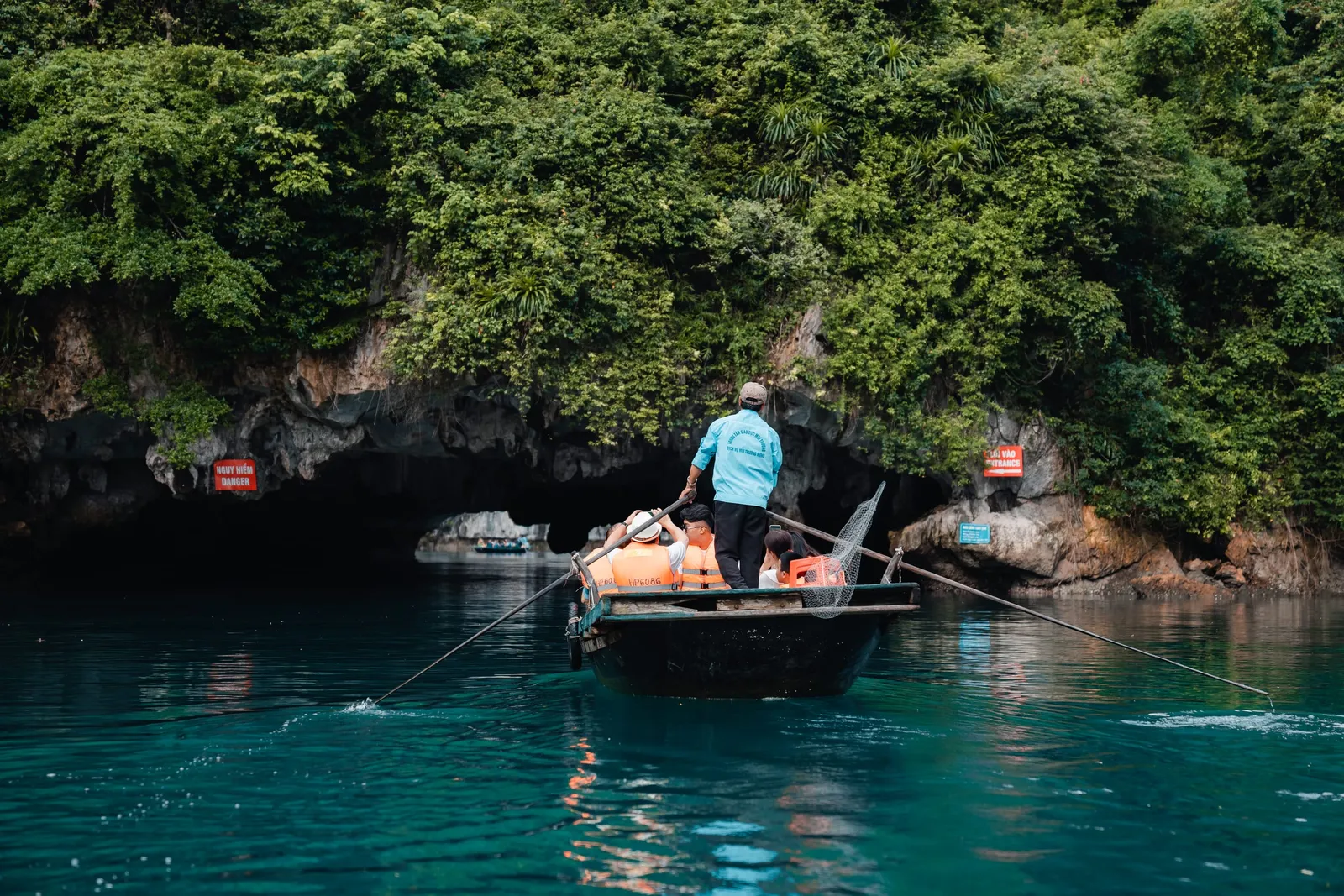 A person in a turquoise shirt rows a small wooden boat with orange life jackets past a cave entrance in green karst cliffs with red signboards mounted on the rock.