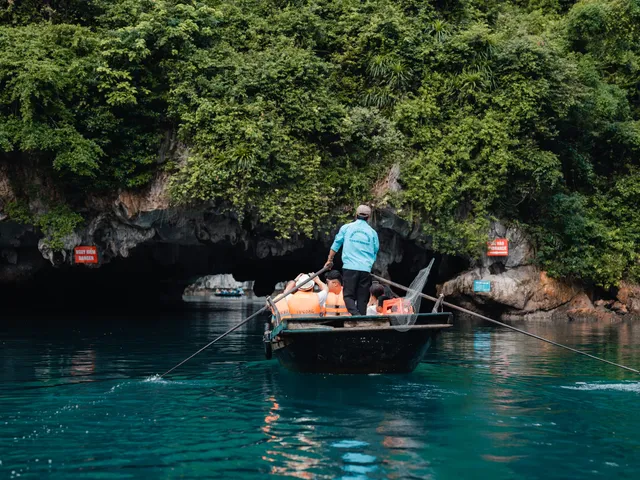 A person in a turquoise shirt rows a small wooden boat with orange life jackets past a cave entrance in green karst cliffs with red signboards mounted on the rock.