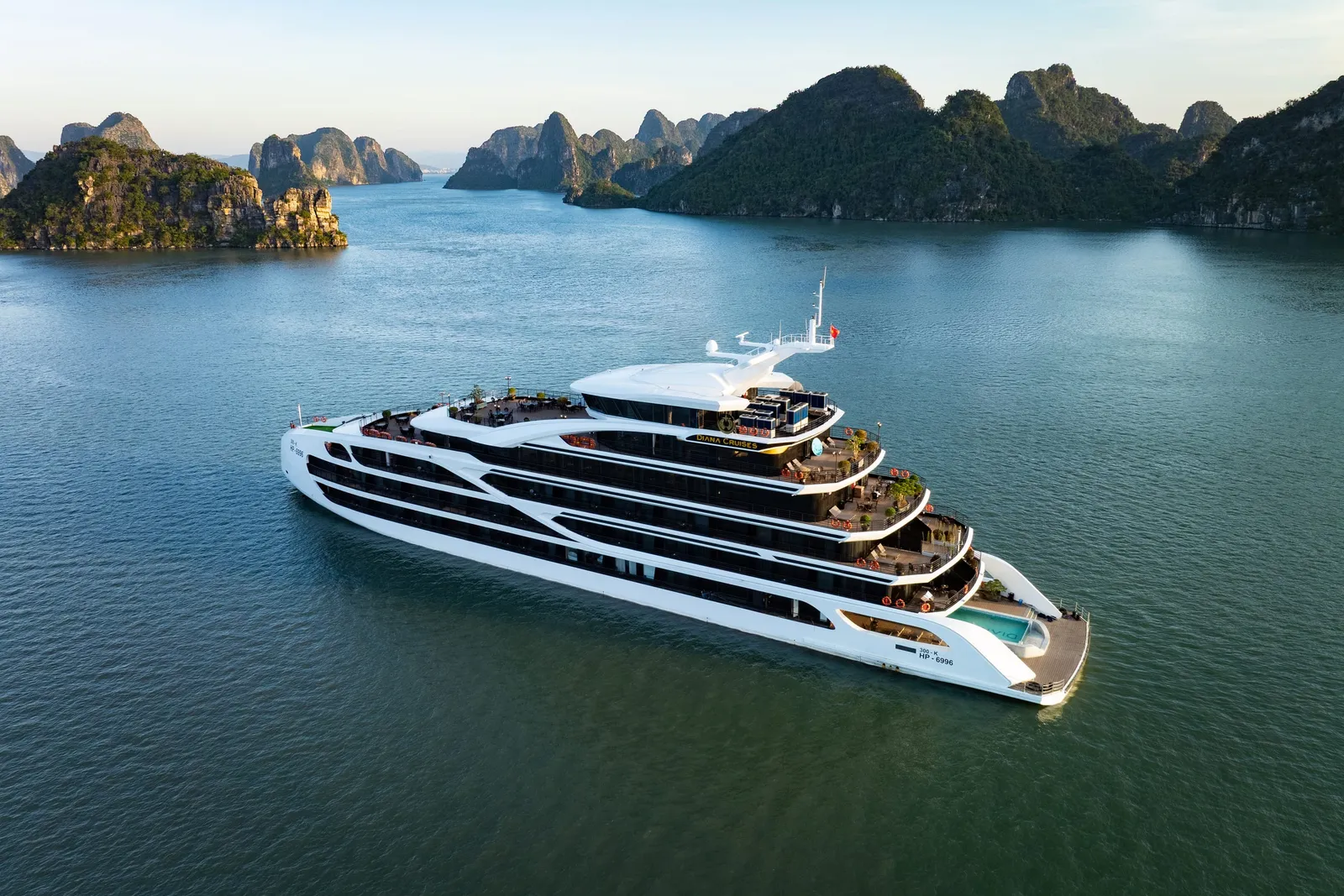 Aerial view of a white multi-deck cruise ship with a pool on the lower deck, sailing among limestone karst islands in calm green-blue water.