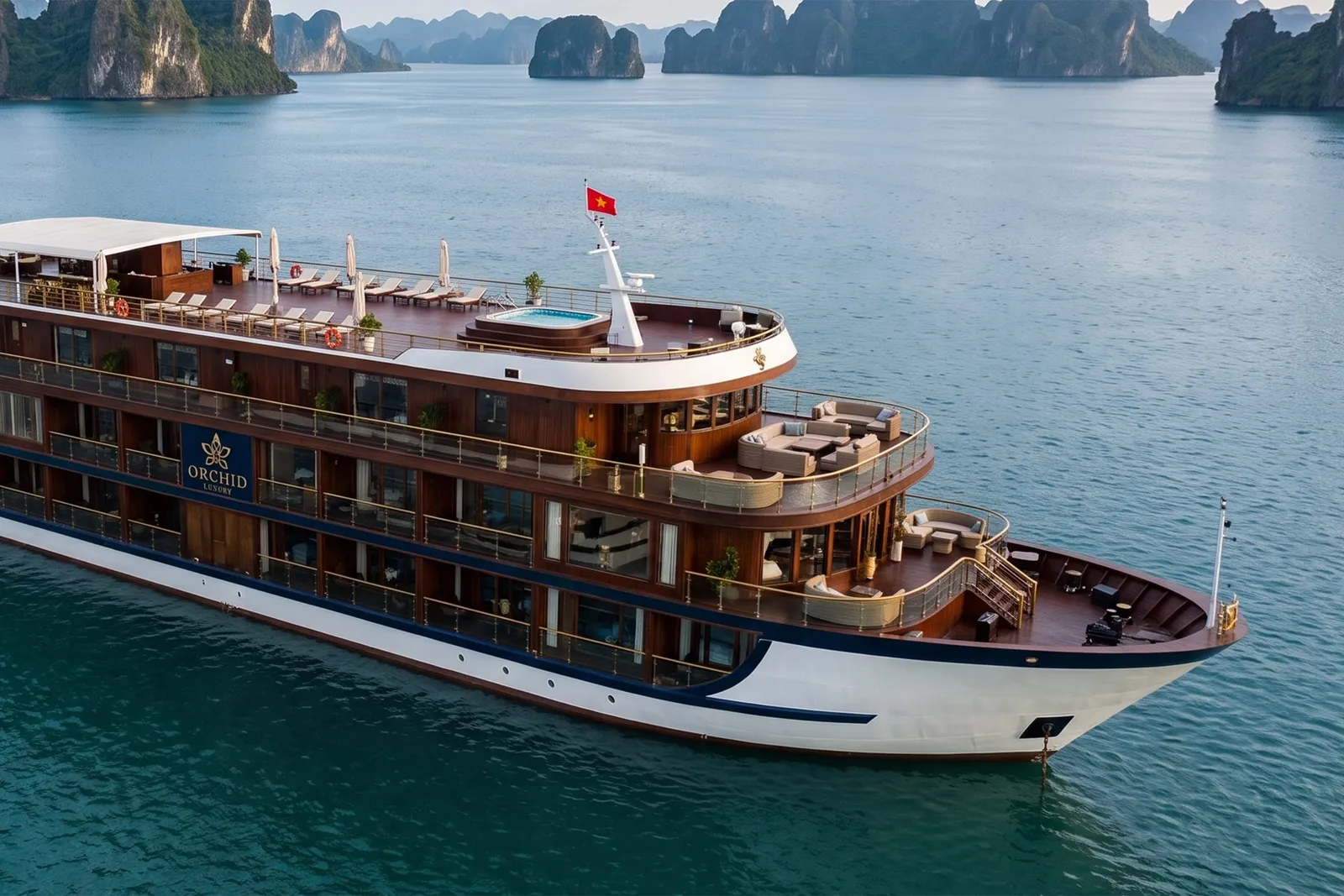A multi-deck cruise ship with brown wood and white hull bearing the name 'ORCHID' on its side floats in calm turquoise water surrounded by dramatic limestone karst formations.