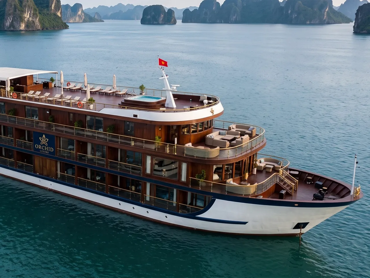 A multi-deck cruise ship with brown wood and white hull bearing the name 'ORCHID' on its side floats in calm turquoise water surrounded by dramatic limestone karst formations.