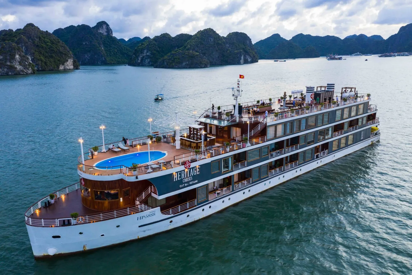 An aerial view of a white and wood-trimmed cruise ship with a blue pool on deck sailing in calm waters surrounded by limestone karst islands.