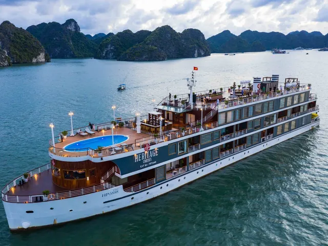An aerial view of a white and wood-trimmed cruise ship with a blue pool on deck sailing in calm waters surrounded by limestone karst islands.