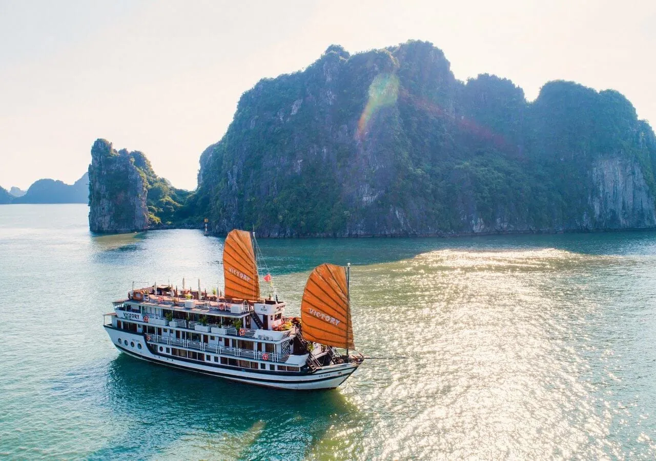 A white multi-deck junk-style cruise vessel with two large orange sails bearing the name 'Victory' sails on calm green water in front of dramatic karst limestone islands.