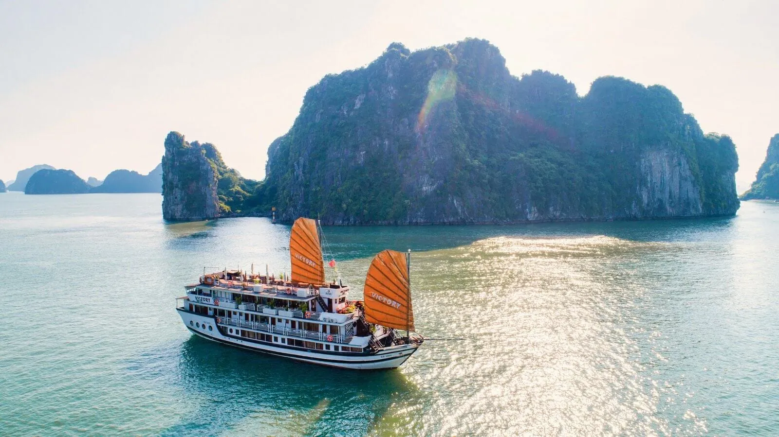 A white multi-deck junk-style cruise vessel with two large orange sails bearing the name 'Victory' sails on calm green water in front of dramatic karst limestone islands.