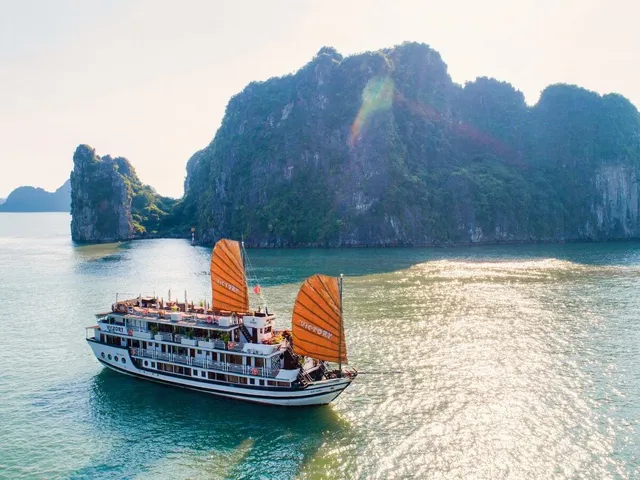 A white multi-deck junk-style cruise vessel with two large orange sails bearing the name 'Victory' sails on calm green water in front of dramatic karst limestone islands.