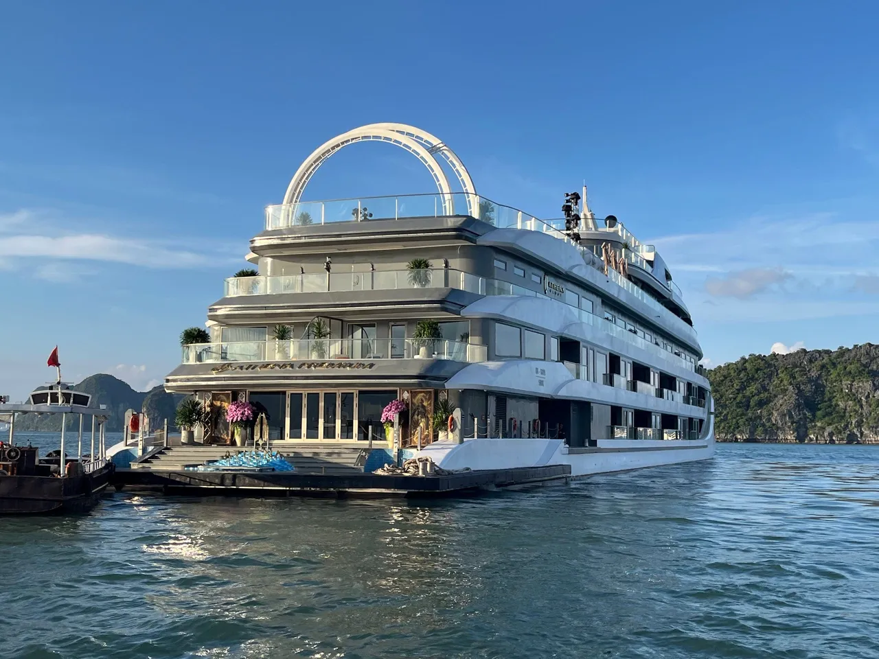 A large multi-deck white cruise ship with an arched structure on its top deck sits on calm water near limestone karst hills, with gold lettering on the hull above the entrance platform and a smaller boat with a red flag at left.