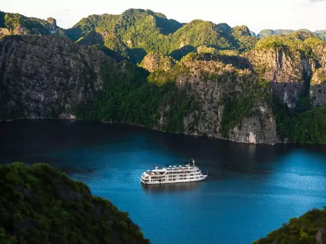 A white multi-deck cruise ship sits in calm blue water surrounded by tall limestone karst formations covered in green vegetation.