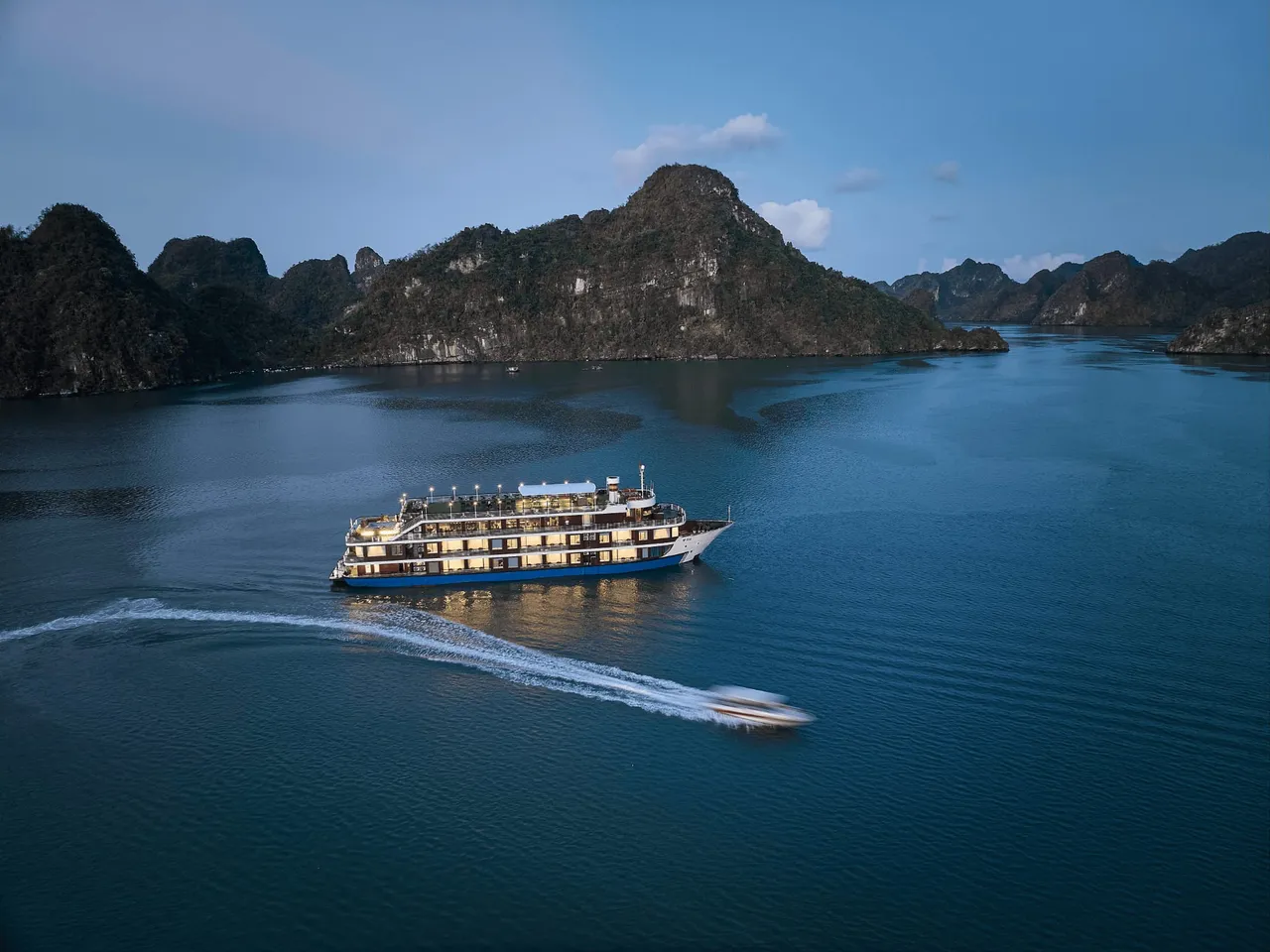 An aerial dusk view of a multi-deck cruise ship with lit windows on calm blue water among forested limestone karst islands, with a speedboat leaving a white wake in the foreground.