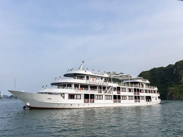 A white multi-deck cruise ship with 'ATHENA CRUISE' painted on its hull sails in calm waters surrounded by limestone karst formations.