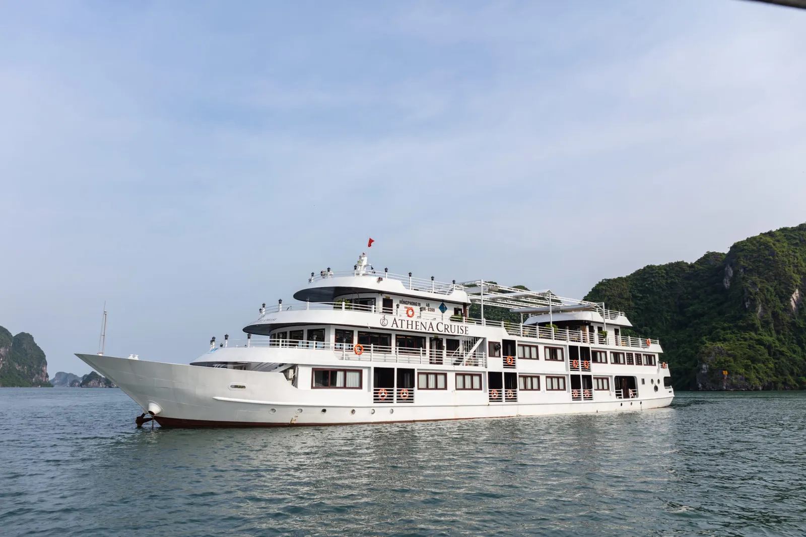 A white multi-deck cruise ship with 'ATHENA CRUISE' painted on its hull sails in calm waters surrounded by limestone karst formations.