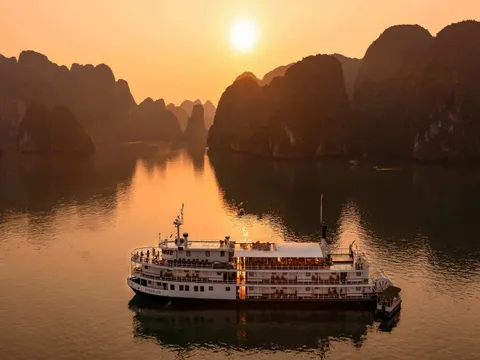 An aerial view at sunset of a white multi-deck paddle-style cruise boat with the name 'Emeraude' on its hull, sailing on calm water among dark limestone karst peaks under an orange sky and low sun.
