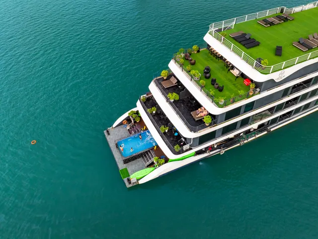 Aerial drone shot of a multi-deck cruise ship with a rooftop grass deck, sunbeds, potted plants, a bow swimming pool, and a green water slide, floating on turquoise water.