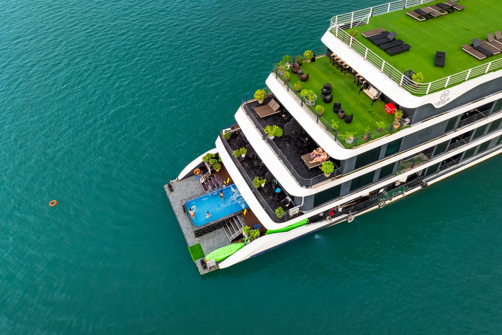 Aerial drone shot of a multi-deck cruise ship with a rooftop grass deck, sunbeds, potted plants, a bow swimming pool, and a green water slide, floating on turquoise water.