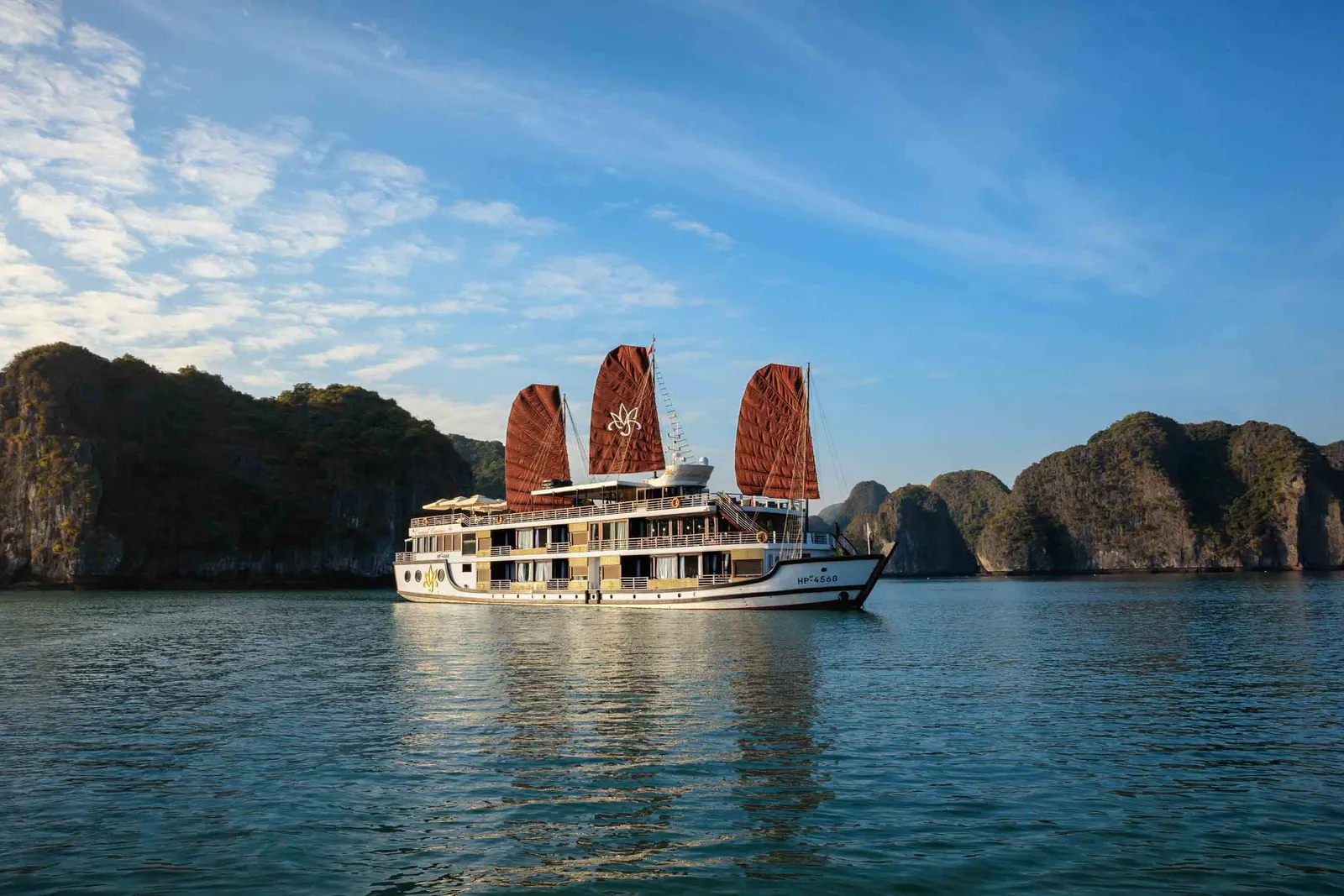 A multi-deck cruise ship with distinctive red sails sits in calm water surrounded by dramatic limestone karst formations under blue sky.