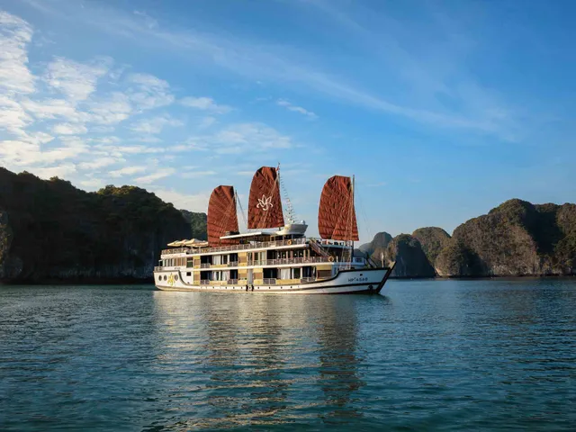 A multi-deck cruise ship with distinctive red sails sits in calm water surrounded by dramatic limestone karst formations under blue sky.