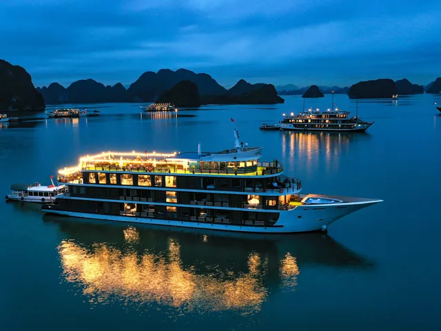 An illuminated multi-deck cruise boat at dusk in a bay surrounded by limestone karst islands with other lit boats in the distance.