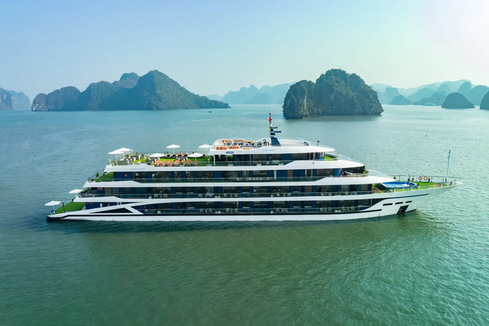 A large white and blue multi-deck cruise ship with green accents sailing in calm turquoise water with limestone karst islands in the background.