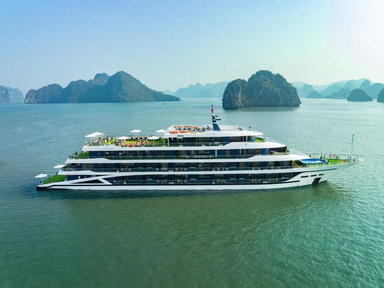A large white and blue multi-deck cruise ship with green accents sailing in calm turquoise water with limestone karst islands in the background.