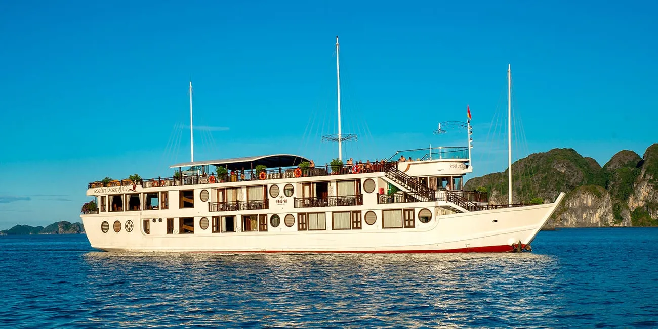 A white cruise boat with multiple decks and circular windows floats on calm blue water with limestone karst formations in the background.