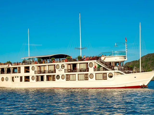 A white cruise boat with multiple decks and circular windows floats on calm blue water with limestone karst formations in the background.