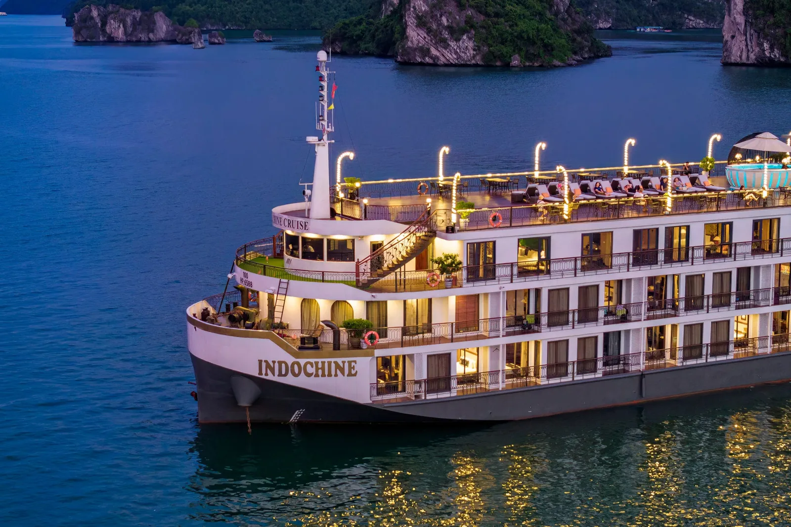 Indochine at dusk with her lights on, the ship name along the white hull and karst islands behind