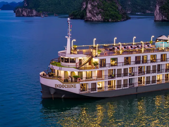 Indochine at dusk with her lights on, the ship name along the white hull and karst islands behind