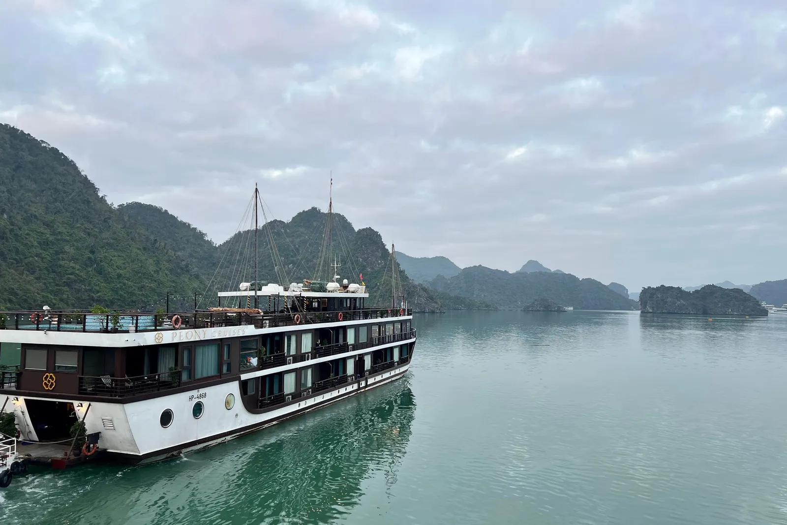 Peony under way on flat water, a white hull under dark upper decks with karst islands crowding the channel behind