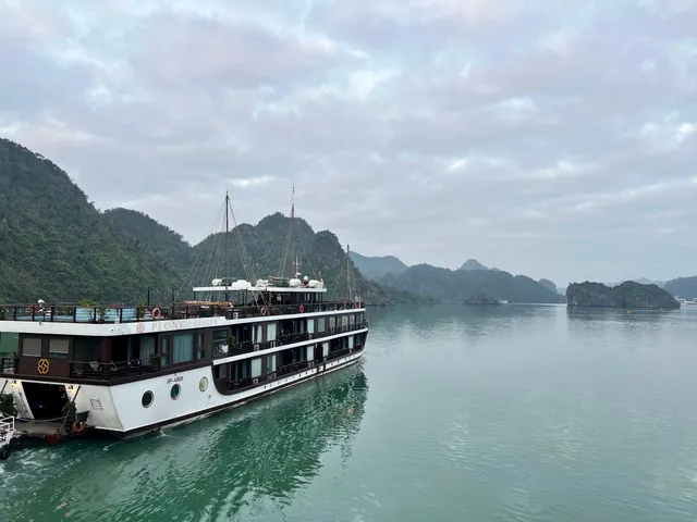 Peony under way on flat water, a white hull under dark upper decks with karst islands crowding the channel behind