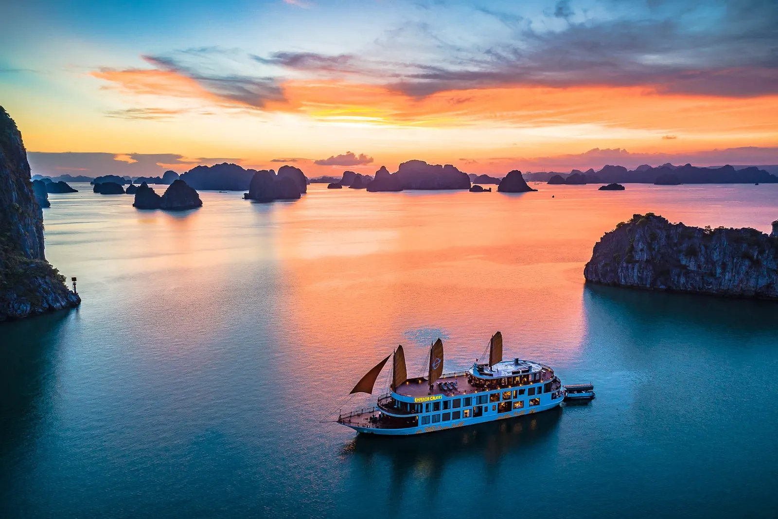 An aerial view at sunset of a blue-hulled cruise ship with orange sails and a lit-up yellow name sign on its hull, floating in calm water among limestone karst islands under an orange and teal sky.