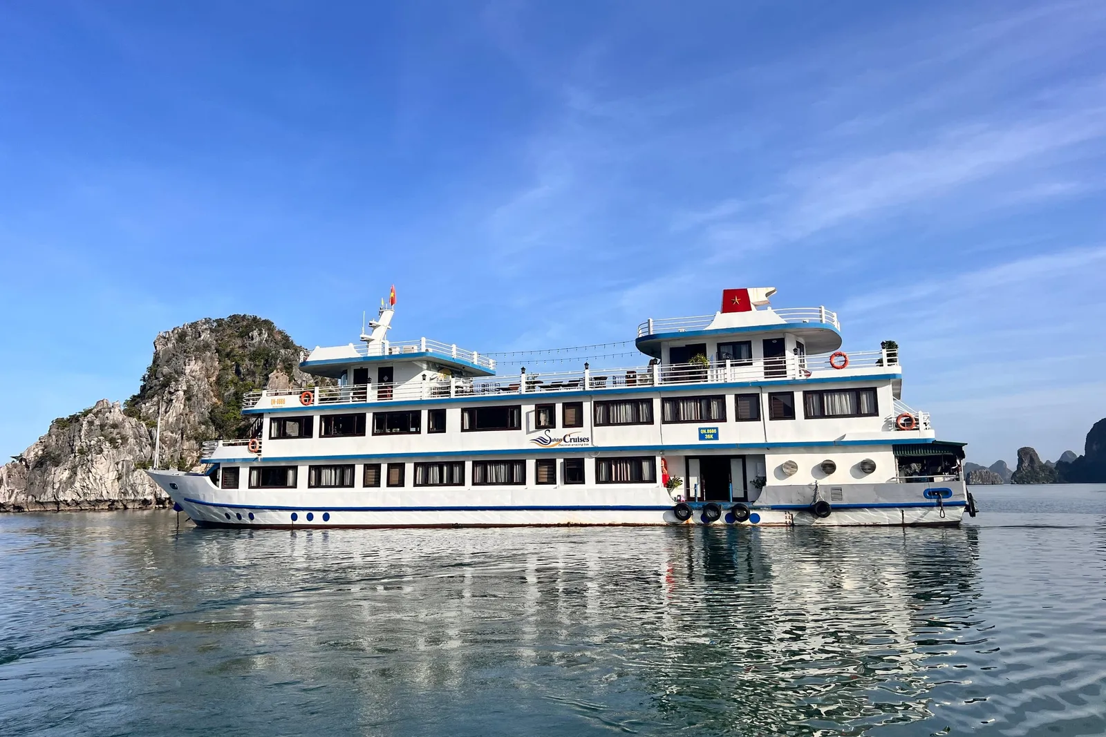 Swan broadside on flat water, a white three-deck hull with blue trim and karst islands close behind