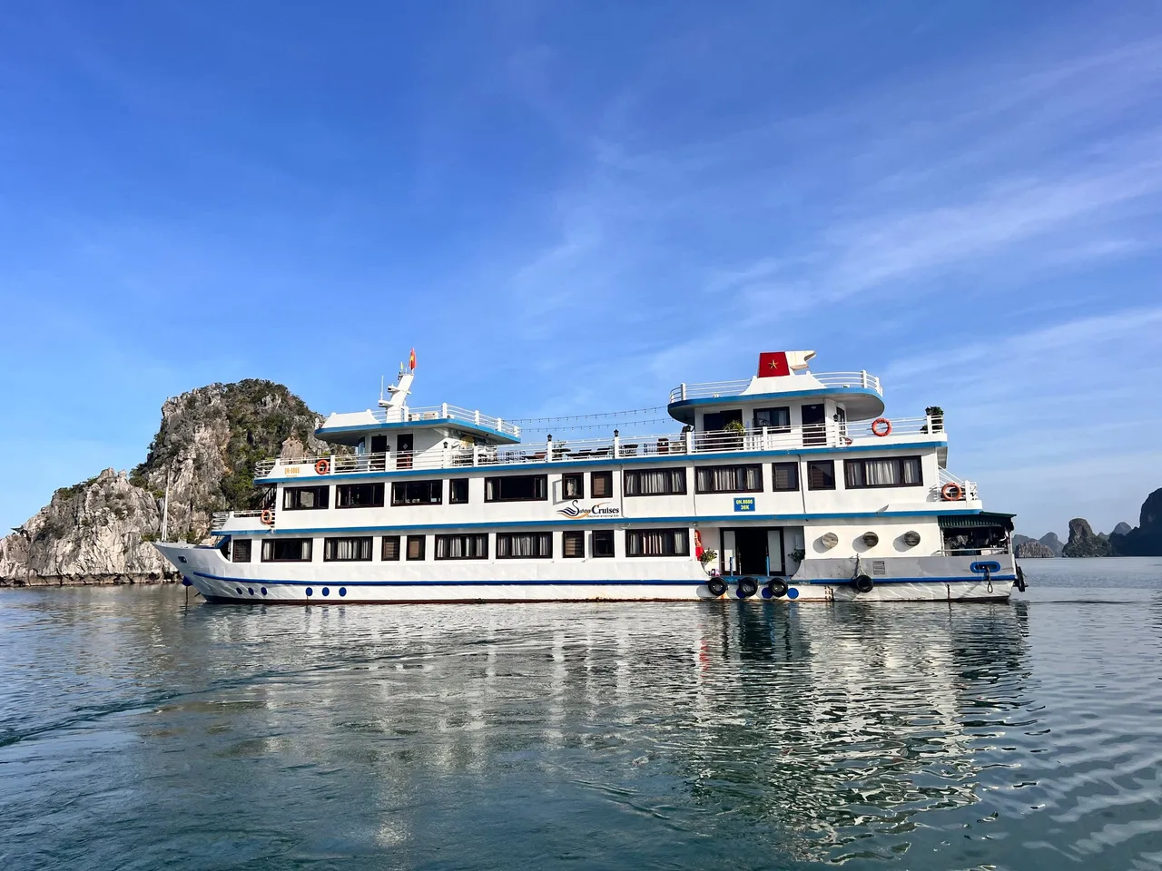 Swan broadside on flat water, a white three-deck hull with blue trim and karst islands close behind