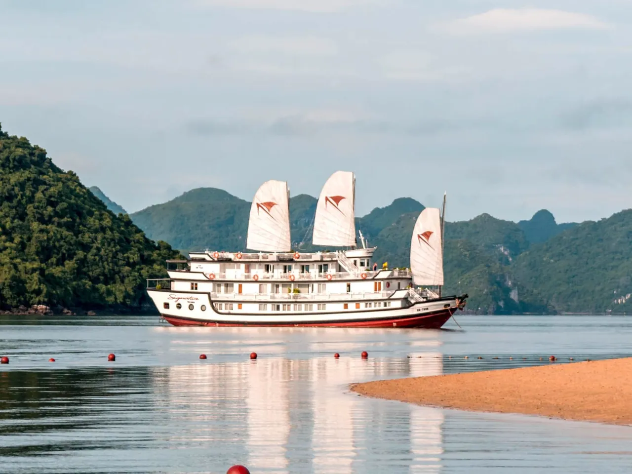 A white multi-deck junk-style cruise ship named 'Signature' with three white sails bearing red logo marks, anchored on calm water surrounded by karst limestone islands, with a sandy shore in the foreground.