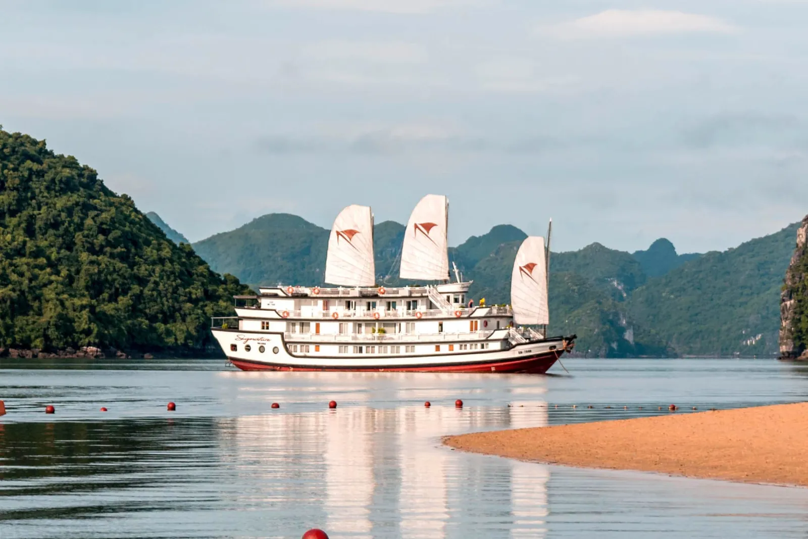 A white multi-deck junk-style cruise ship named 'Signature' with three white sails bearing red logo marks, anchored on calm water surrounded by karst limestone islands, with a sandy shore in the foreground.