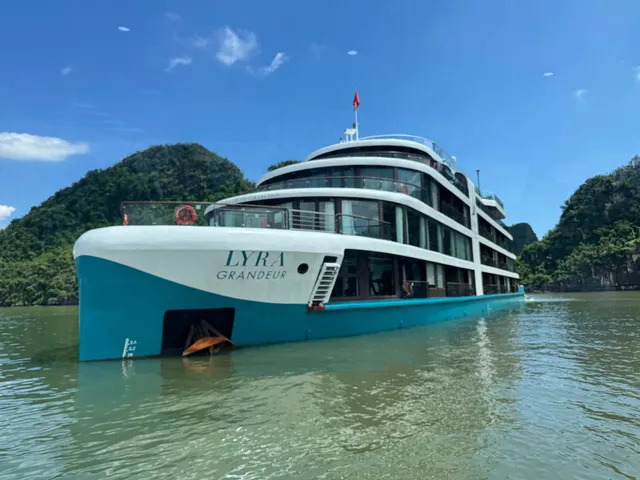 Lyra Grandeur broadside close to a green island, the ship name along a teal hull below three white decks