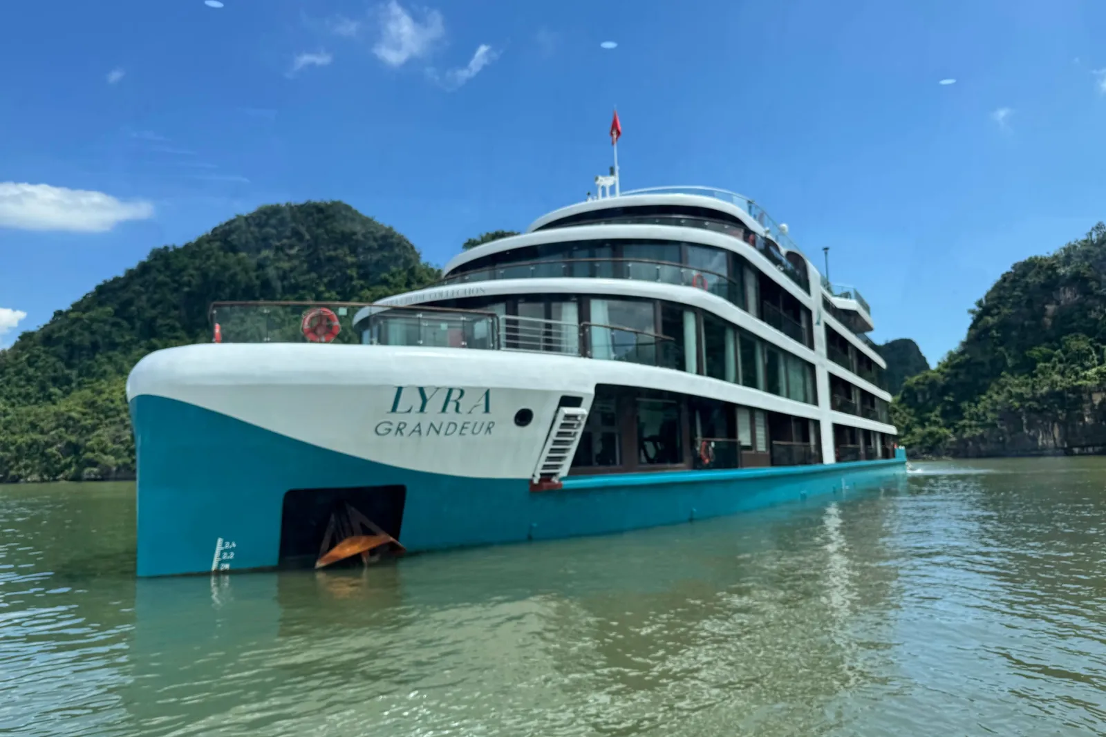 Lyra Grandeur broadside close to a green island, the ship name along a teal hull below three white decks