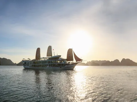A white multi-deck cruise boat with four sails and a small red flag sails on calm water toward a low sun, with limestone karst islands in the hazy background; small text is visible on the hull but no overlay.
