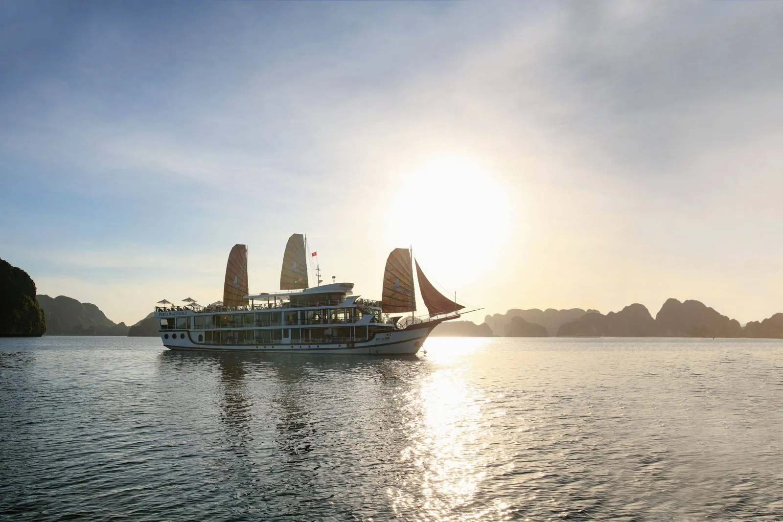 A white multi-deck cruise boat with four sails and a small red flag sails on calm water toward a low sun, with limestone karst islands in the hazy background; small text is visible on the hull but no overlay.