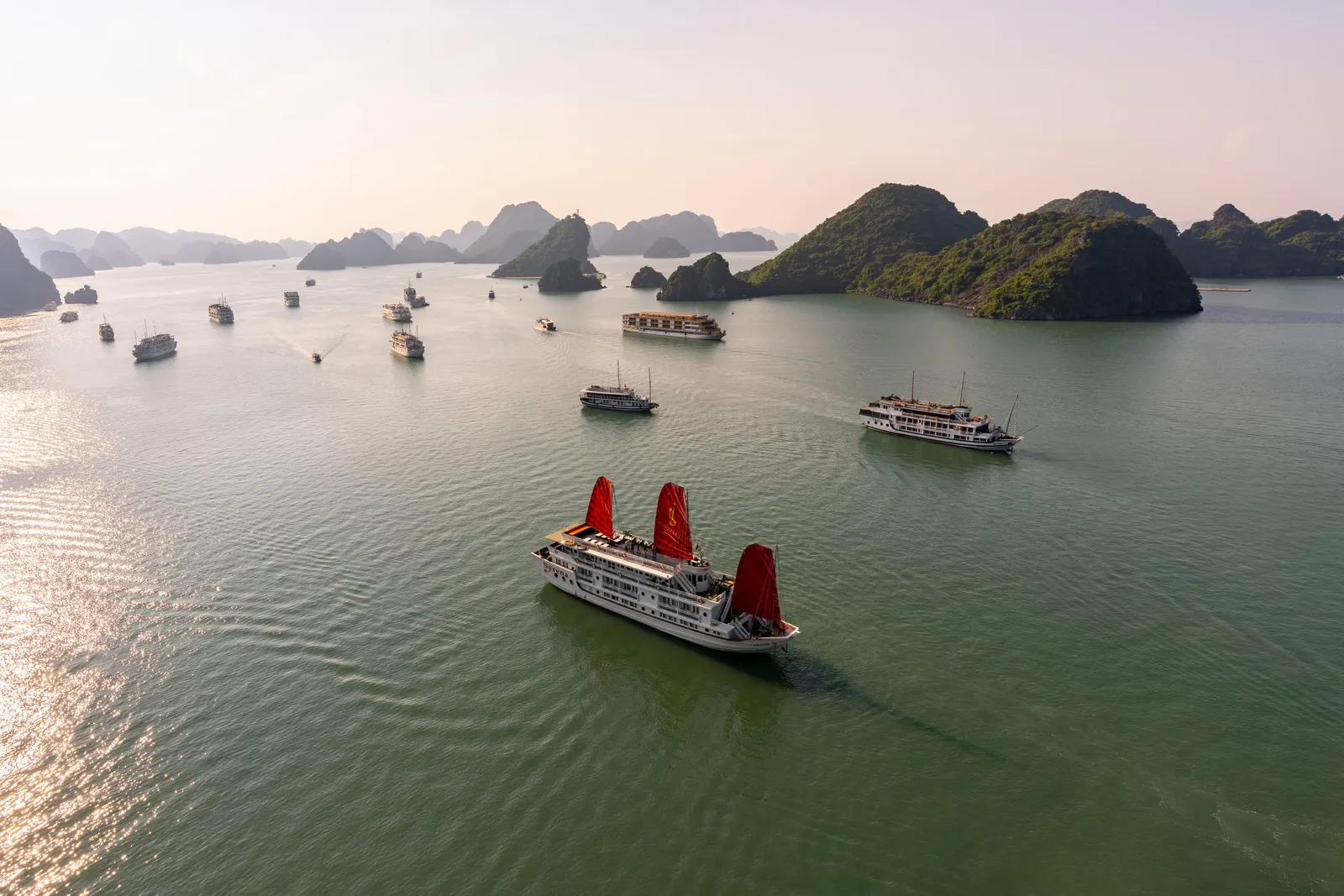 Aerial view of a white cruise boat with three red sails in calm green water, with other cruise boats and forested limestone karst islands in the hazy background.