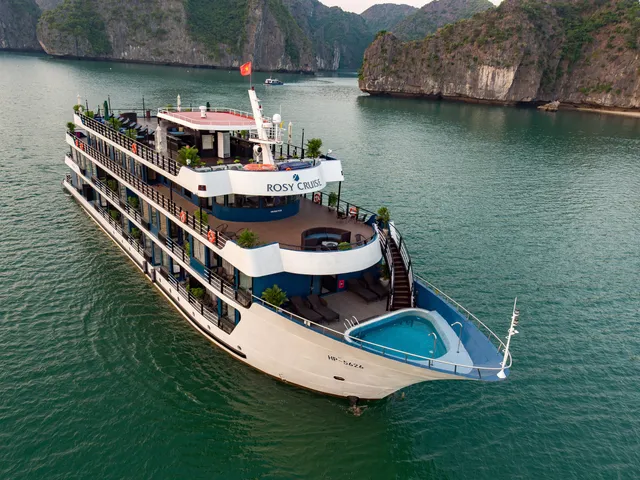 Rosy Cruise broadside on flat green water, four white decks with a plunge pool set into the bow, karst cliffs rising close behind