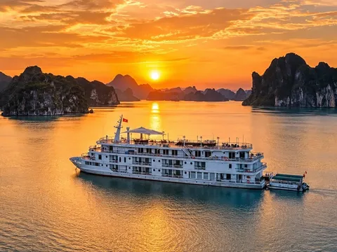 An aerial view of a white multi-deck cruise boat with a small tender alongside, on calm water among forested limestone karst islands at sunset.