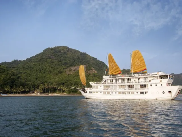 A white multi-deck cruise boat with golden-orange junk sails and a circular emblem on the sails sits on calm water in front of a forested hill with palm trees and a small beach.