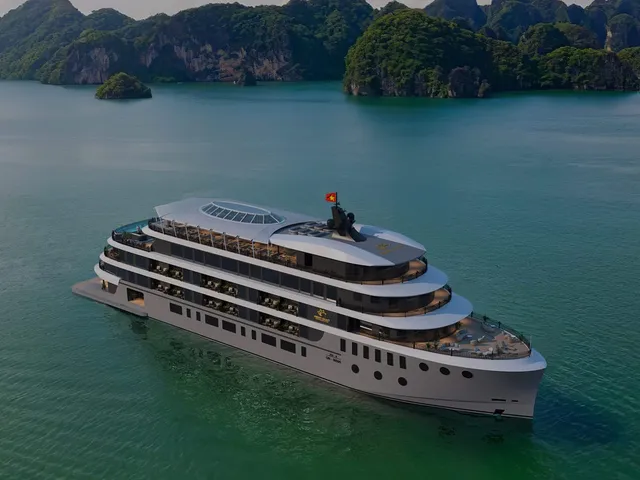 An aerial view of a large multi-deck white cruise ship with a Vietnamese flag on calm green water, with forested limestone karst islands in the background and a small logo emblem on the vessel's side.