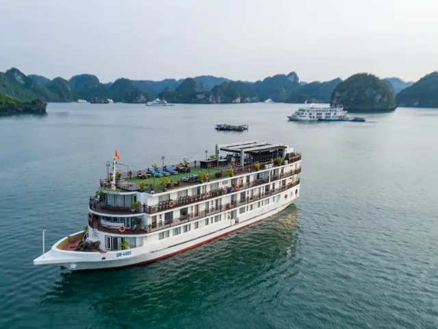 An aerial view of a multi-deck white cruise ship with a registration number on its hull, sailing on calm green water among tree-covered limestone islands with another ship and a small boat in the distance.