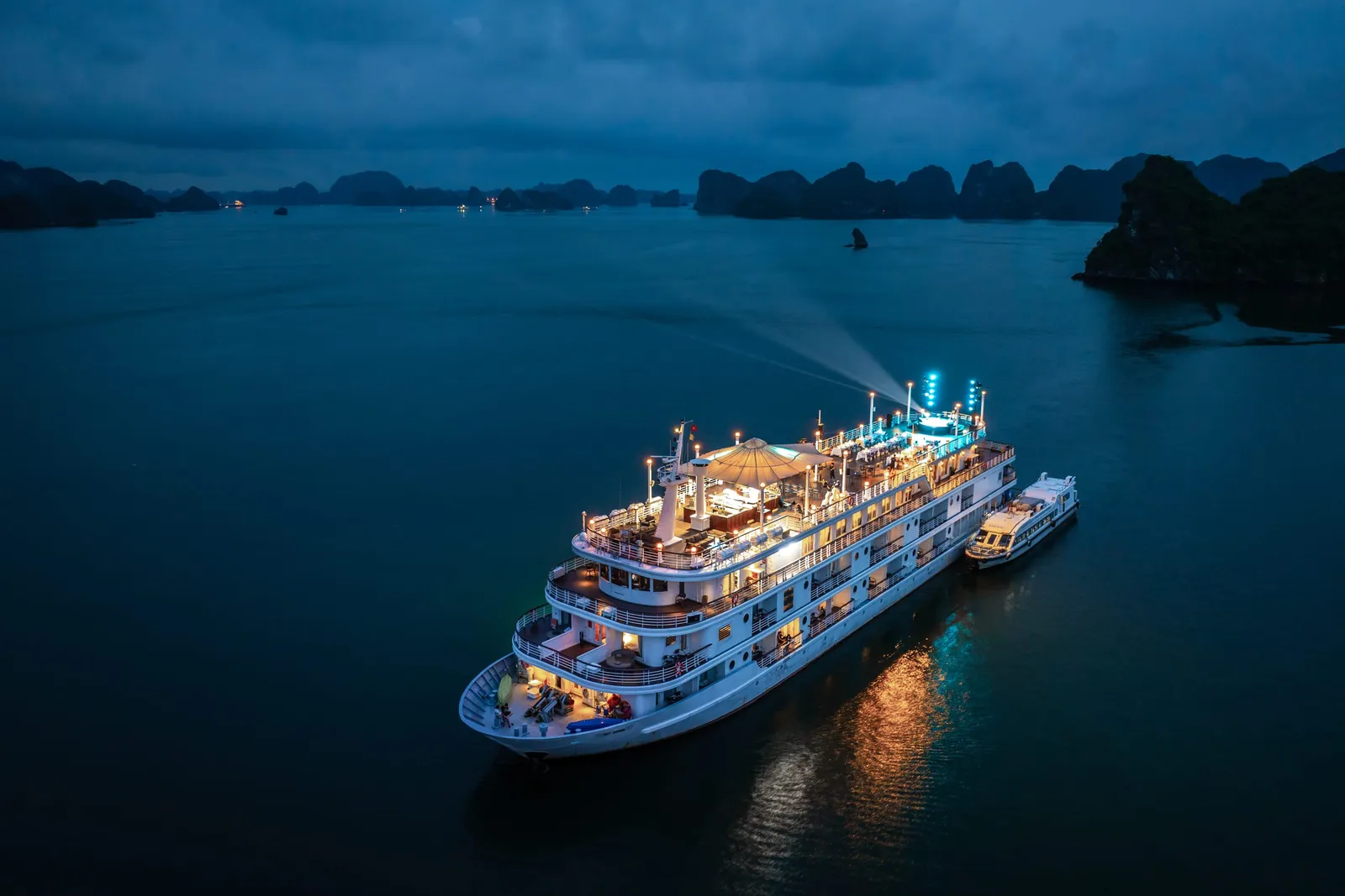 An illuminated multi-deck cruise ship sits in calm water at dusk surrounded by limestone karst formations characteristic of Ha Long Bay.