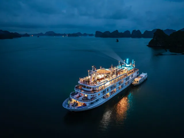 An illuminated multi-deck cruise ship sits in calm water at dusk surrounded by limestone karst formations characteristic of Ha Long Bay.